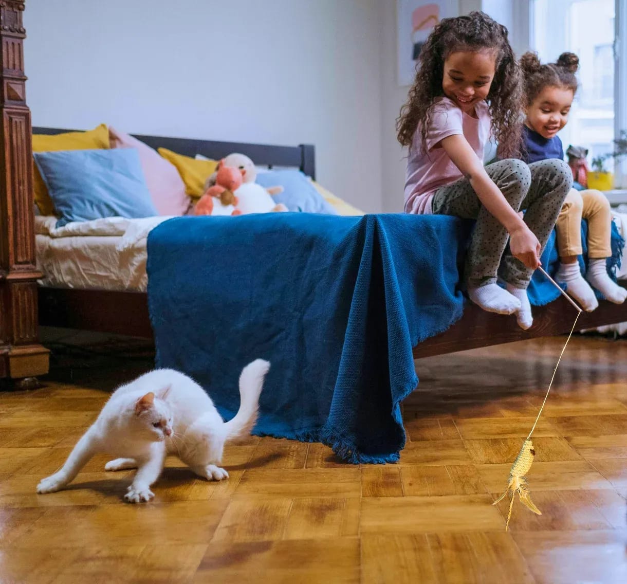 Two children playing with a white cat using a sisal cat teaser toy on a bed.